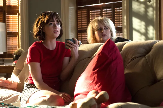 Two women sitting on a couch, one holding a mug, with sunlight streaming through window blinds.