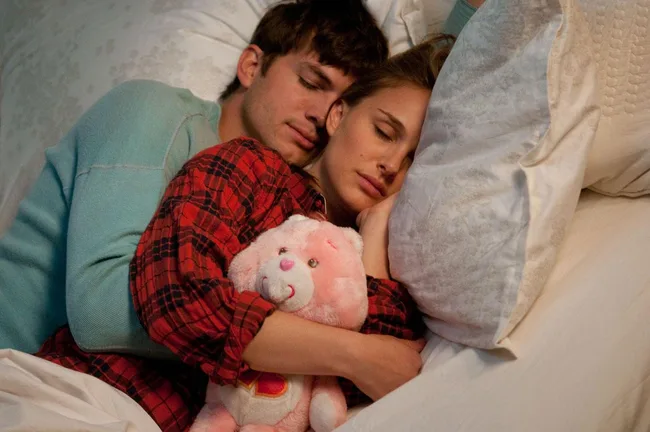 A couple cuddles while sleeping, the woman holding a pink teddy bear, both on white pillows in a serene setting.