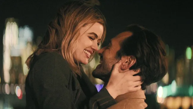 A couple embracing and smiling at night with a city skyline in the background.