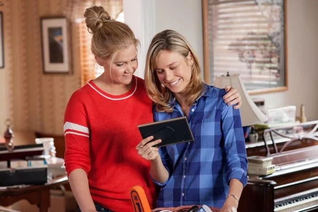 Two women smiling and looking at a photo frame, standing in a cozy room with a piano and table in the background.