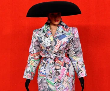 A model in a large black hat and a vibrant collage-printed coat stands before a red backdrop at Balenciaga SS 2022.