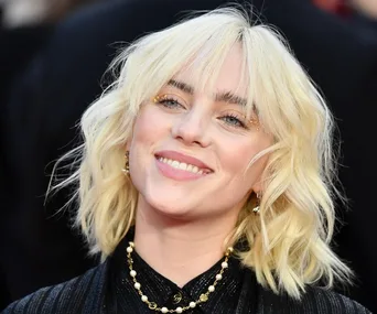 A person with blonde hair smiles, wearing a black outfit and pearl necklace.