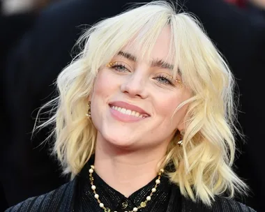 A person with blonde hair smiles, wearing a black outfit and pearl necklace.