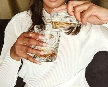 A person in a white shirt pours whiskey from a small bottle into a glass, wearing several gold rings.