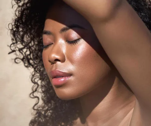 Person with curly hair basks in sunlight, highlighting smooth, glowing skin and closed eyes.