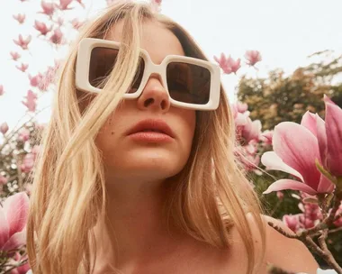 Woman wearing large white sunglasses amidst blooming pink flowers.