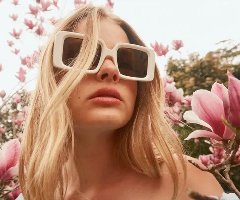 Woman wearing large white sunglasses amidst blooming pink flowers.