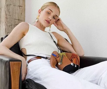 Woman in a sleeveless top and white pants, seated with a colorful purse, leaning on a chair.