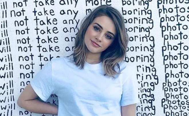 Actress in white t-shirt stands against a wall with repeated text, "I will not take any more boring photos."