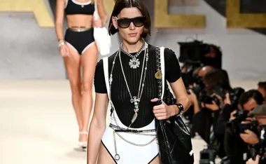 A model in black and white swimwear, layered jewelry, and sunglasses walks the runway at Chanel Spring/Summer 2022.