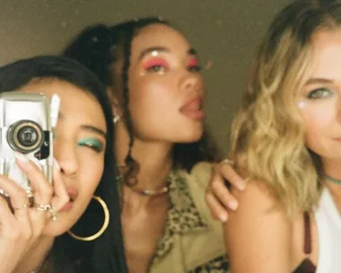 Three women taking a selfie with a vintage camera, showing vibrant makeup and fashion.