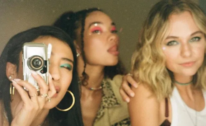 Three women taking a selfie with a vintage camera, showing vibrant makeup and fashion.