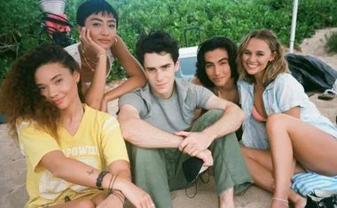 Group of five young adults smiling while sitting on a beach, surrounded by greenery.