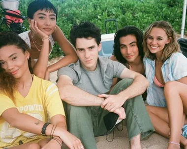 Group of five young adults smiling while sitting on a beach, surrounded by greenery.