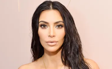 A woman with dark, wavy hair poses against a light background, wearing neutral makeup and a thin necklace.