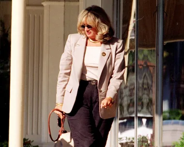 A woman in sunglasses and business attire walks outside, holding a bag, with reflections visible in a glass storefront.