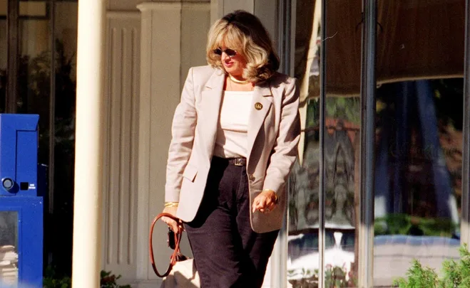 A woman in sunglasses and business attire walks outside, holding a bag, with reflections visible in a glass storefront.