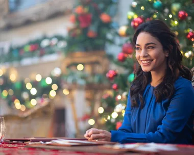 Woman in blue dress smiling, seated in festive room with decorated Christmas tree; cozy, cheerful atmosphere.