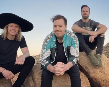 Three men sitting on rocks outdoors, one wearing a large hat, all smiling.