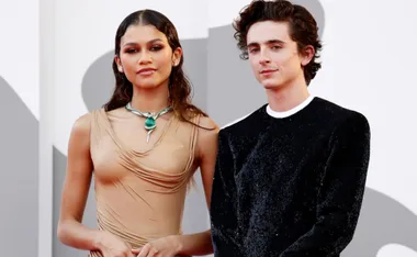 Two actors from the "Dune" movie pose on a red carpet; one in a tan dress, the other in a black suit.