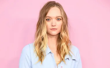 A person with long blonde hair wearing a light blue shirt, set against a pink background.