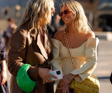 Two stylish women with vibrant bags chatting and smiling in the sunlight.