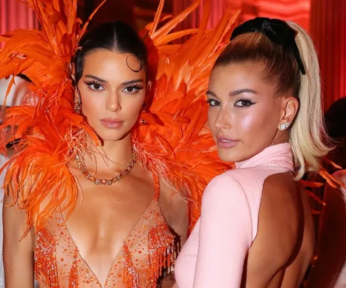 Two women pose at an event, one in an orange feather dress, the other in a pink backless outfit with a ponytail.