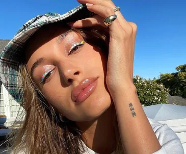 A woman with colorful eye makeup and glossy lips poses in sunlight while wearing a plaid hat.