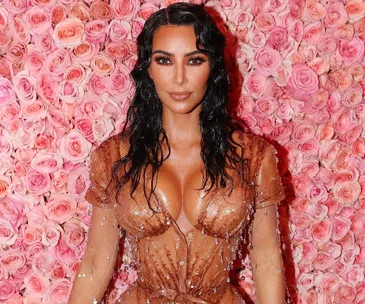 Kim Kardashian in a sheer, beaded dress at an event, standing in front of a pink rose backdrop.