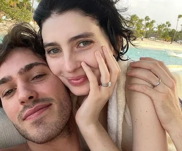 A couple posing together, smiling, with a beach in the background. Both wear wedding rings.