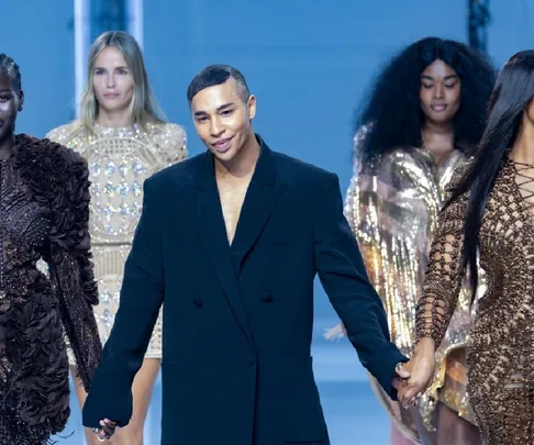 Olivier Rousteing walks down the runway in a black suit, holding hands with models wearing glamorous dresses.