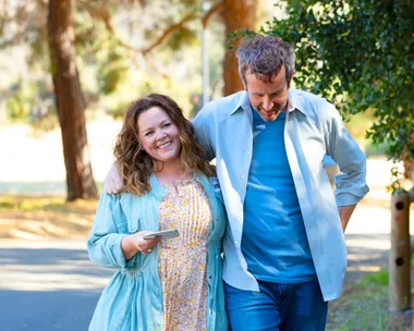 Two people walking closely in a sunny park setting, one smiling while holding a phone.