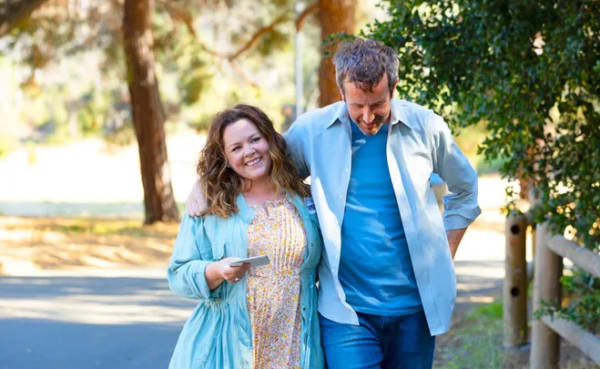 Two people walking closely in a sunny park setting, one smiling while holding a phone.