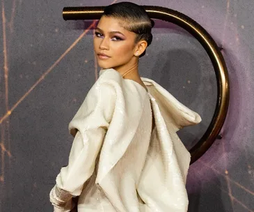 Zendaya in an elegant off-white gown with structured shoulders, posing against a backdrop, at a fashion event.