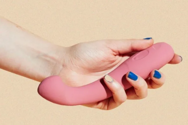 A hand with blue nails holds a pink, curved vibrator against a beige background.