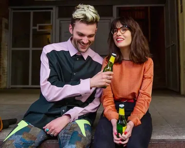 A man and woman sitting and smiling with beer bottles, dressed in colorful casual outfits.