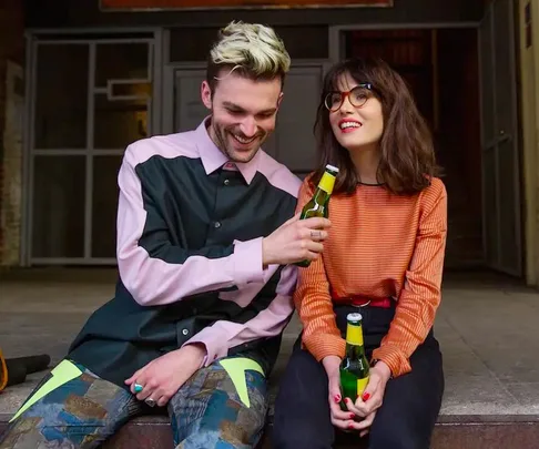 A man and woman sitting and smiling with beer bottles, dressed in colorful casual outfits.