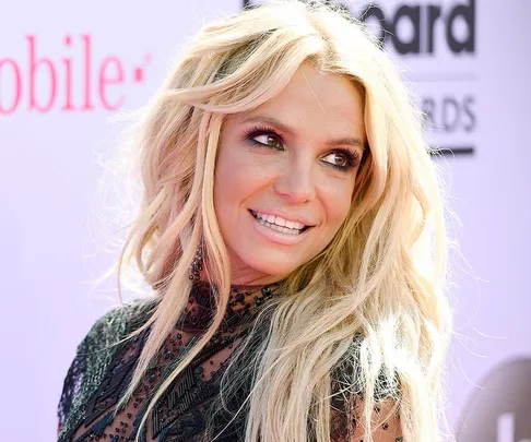 Blonde woman with long hair smiling, wearing a black lace dress at an event with a pink and black sign in the background.