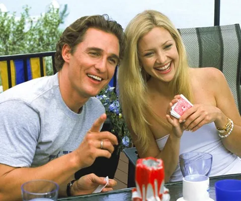 A couple laughing while playing a game with cards outdoors. They sit at a table with drinks and a candle.