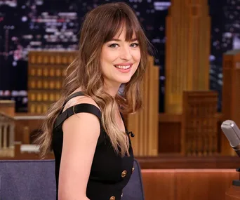 A woman with long brown hair and bangs smiles while seated in a studio setting.