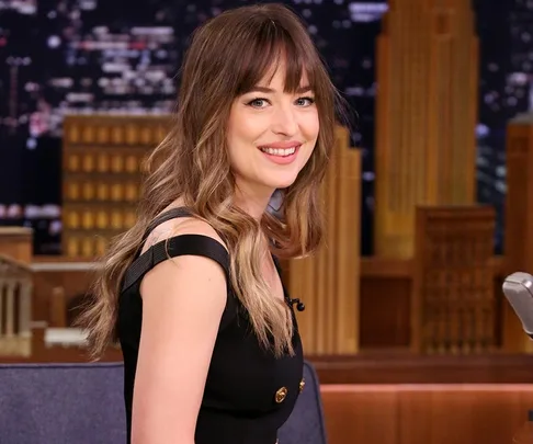 Smiling woman with long hair in black dress on a TV show set background.