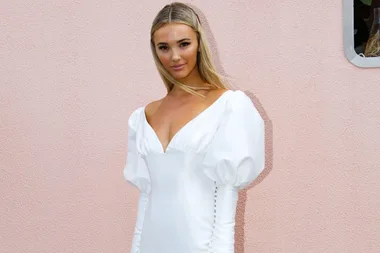 Woman in a white dress with puffed sleeves stands against a light pink wall.