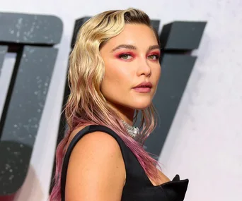 Woman with wavy hair and pink highlights, wearing a black dress and choker, against a light background.