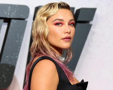 Woman with wavy hair and pink highlights, wearing a black dress and choker, against a light background.