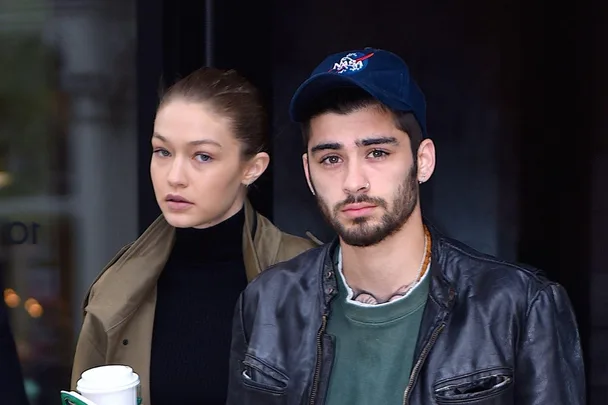 A man and a woman walking together, the man wearing a cap and the woman holding a coffee cup.