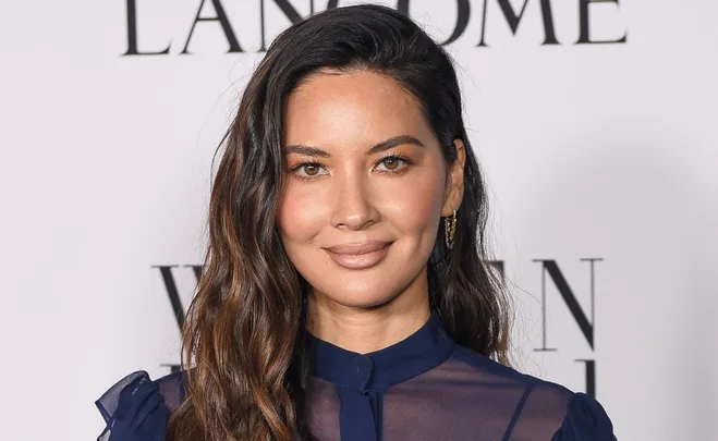 A woman with long, wavy hair and a navy blouse smiles at an event with a blurred text background.