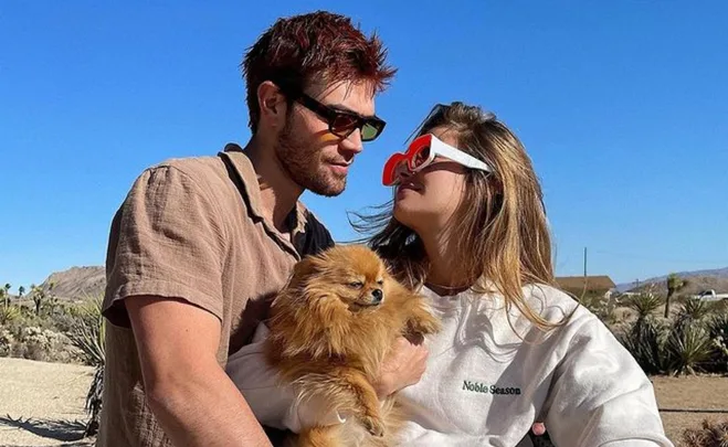 Couple in sunglasses holding a small dog, standing in a sunny desert landscape.