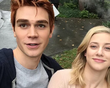 Two actors from "Riverdale" take a selfie outdoors, smiling with greenery in the background.