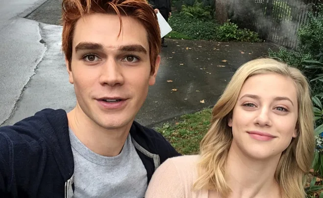 Two actors from "Riverdale" take a selfie outdoors, smiling with greenery in the background.