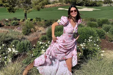 Woman in a pink floral maxi dress, smiling outdoors amid greenery and blooming white flowers.